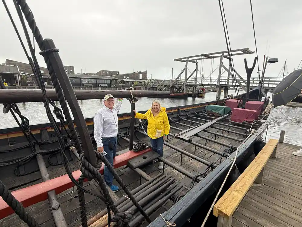 Two people on wooden Viking ship deck by river and bridge